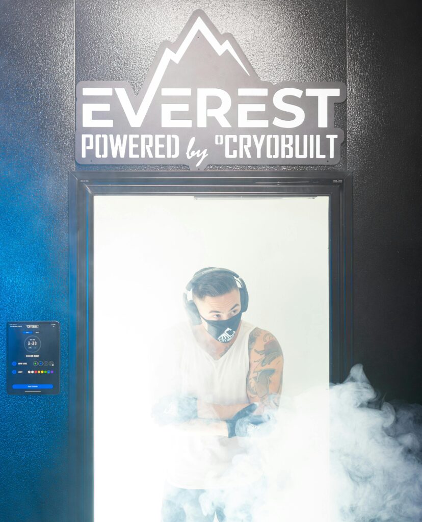 A man wearing gloves, headphones, and a face mask stands inside a cryotherapy chamber surrounded by cold vapor. The sign above reads “Everest Powered by Cryobuilt.”