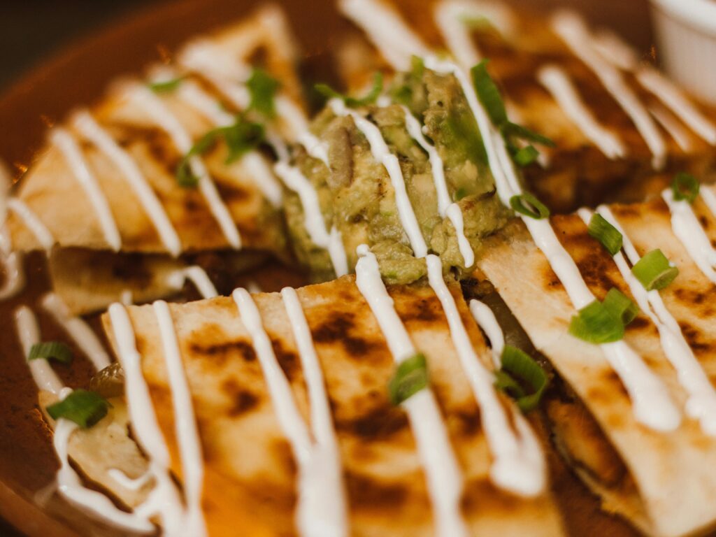 Close-up of sliced quesadillas drizzled with crema, topped with guacamole and chopped green onions on a brown plate.