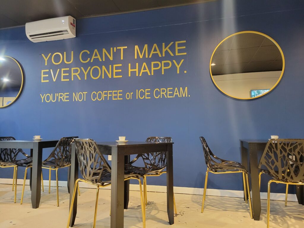 Modern cafe seating area at Grind Central Station in DeQuincy, Louisiana, featuring a bold blue wall mural that reads “You can’t make everyone happy. You’re not coffee or ice cream."