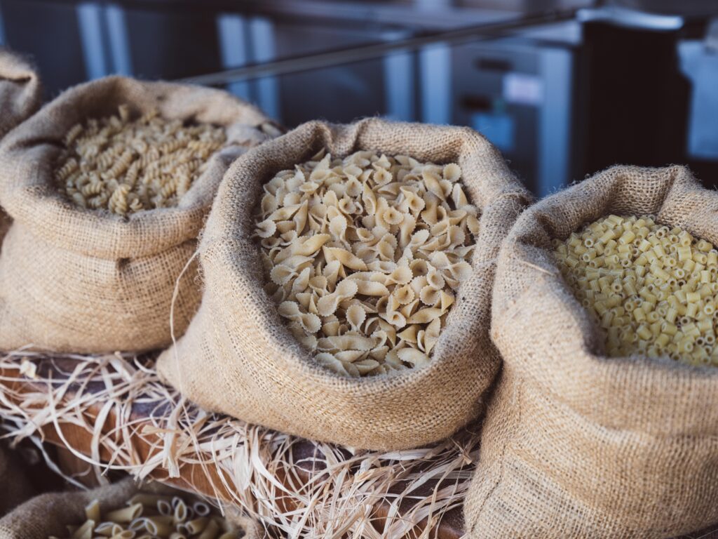 Burlap sacks filled with different shapes of uncooked Italian pasta, displayed in a rustic setting.