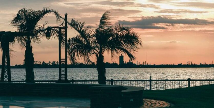Sunset over the Lake Charles lakefront with silhouetted palm trees and a waterfront promenade.