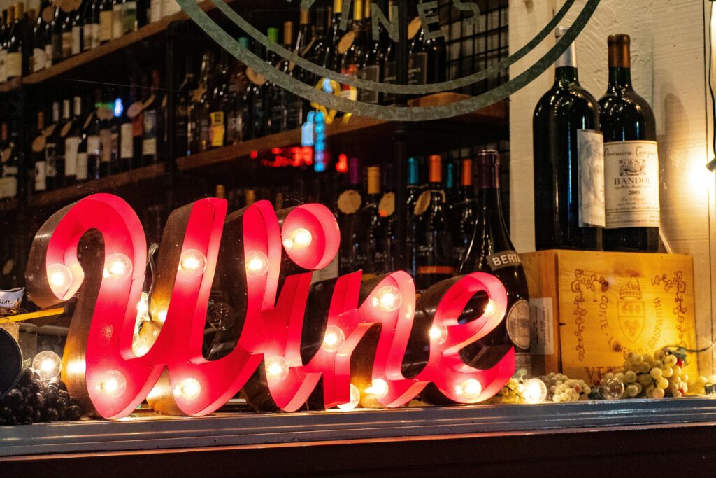 Bright red marquee sign spelling ‘Wine’ lit up in a shop window, surrounded by wine bottles, grape decorations, and wine crates. Perfect for a wine festival or celebration.