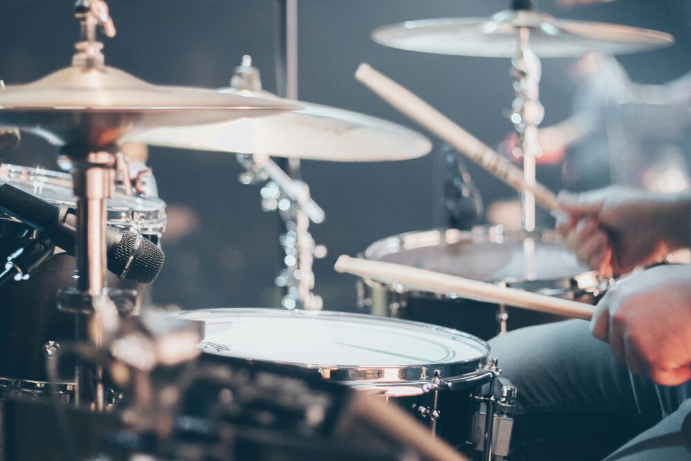 A musician's hands in motion with drumsticks over a black and chrome drum kit, with shiny cymbals and a microphone visible in the stage lighting.