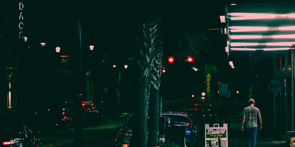 A person stands on a city street or stage at night, dwarfed by large, out-of-focus red and white bokeh lights in the foreground.